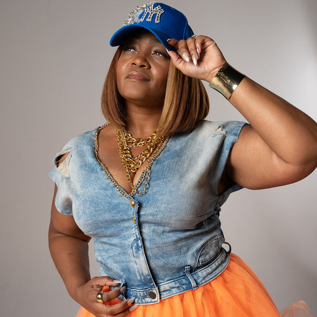 A woman posing in a light blue denim vest, a bright orange tulle skirt, a blue baseball cap, and gold necklaces against a plain gray studio background.