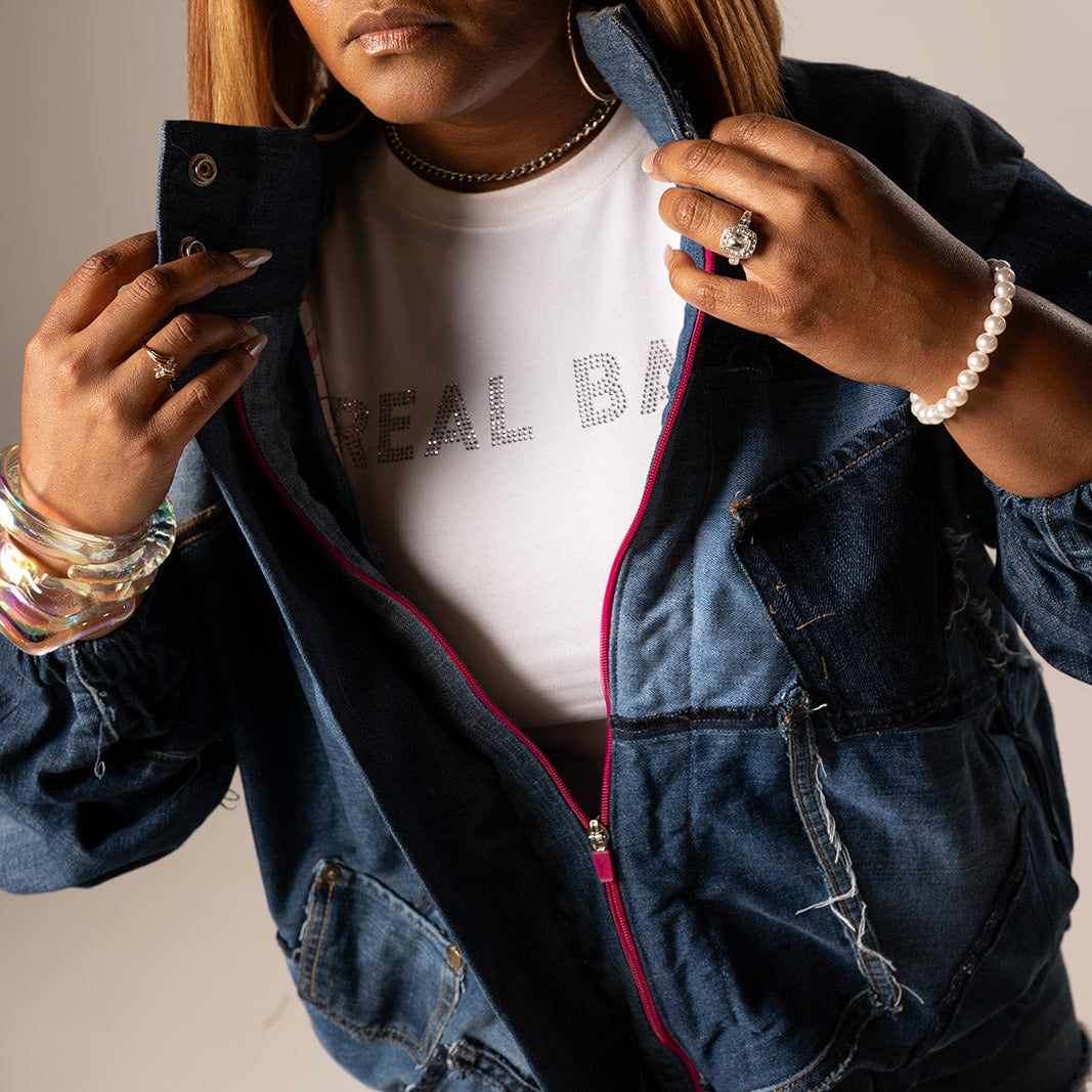 A woman wearing oversized dark sunglasses, a white graphic T-shirt, and a distressed blue denim jacket with a pink zipper, holding a phone in front of her face against a plain beige background.