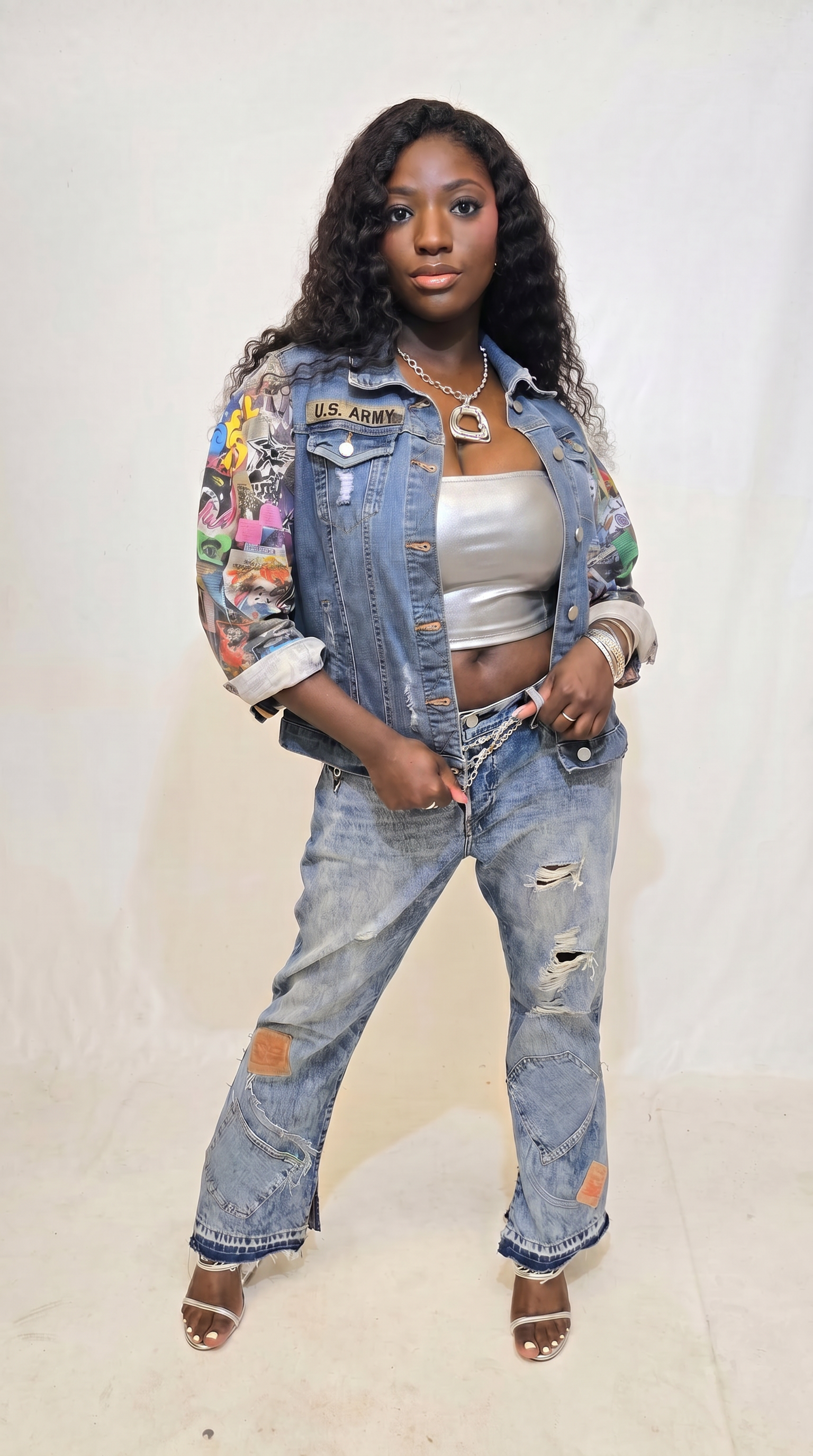 A woman modeling a custom denim outfit consisting of a heavily embellished denim jacket with colorful patches and embroidery, matching distressed denim jeans with ripped patches, a silver crop top, and brown heeled sandals, set against a plain off-white backdrop.