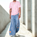 Person wearing a pink tie-dye shirt and blue patchwork palazzo jeans walking in a sunlit concrete area.
