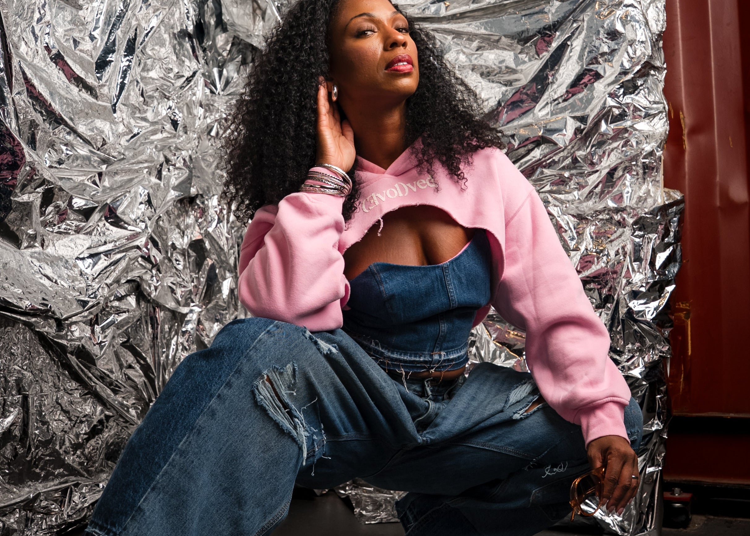 A woman crouches in front of a crumpled silver foil backdrop, wearing a pink cropped hoodie over a blue denim cutout top, wide-leg distressed jeans, and metallic silver high heels.