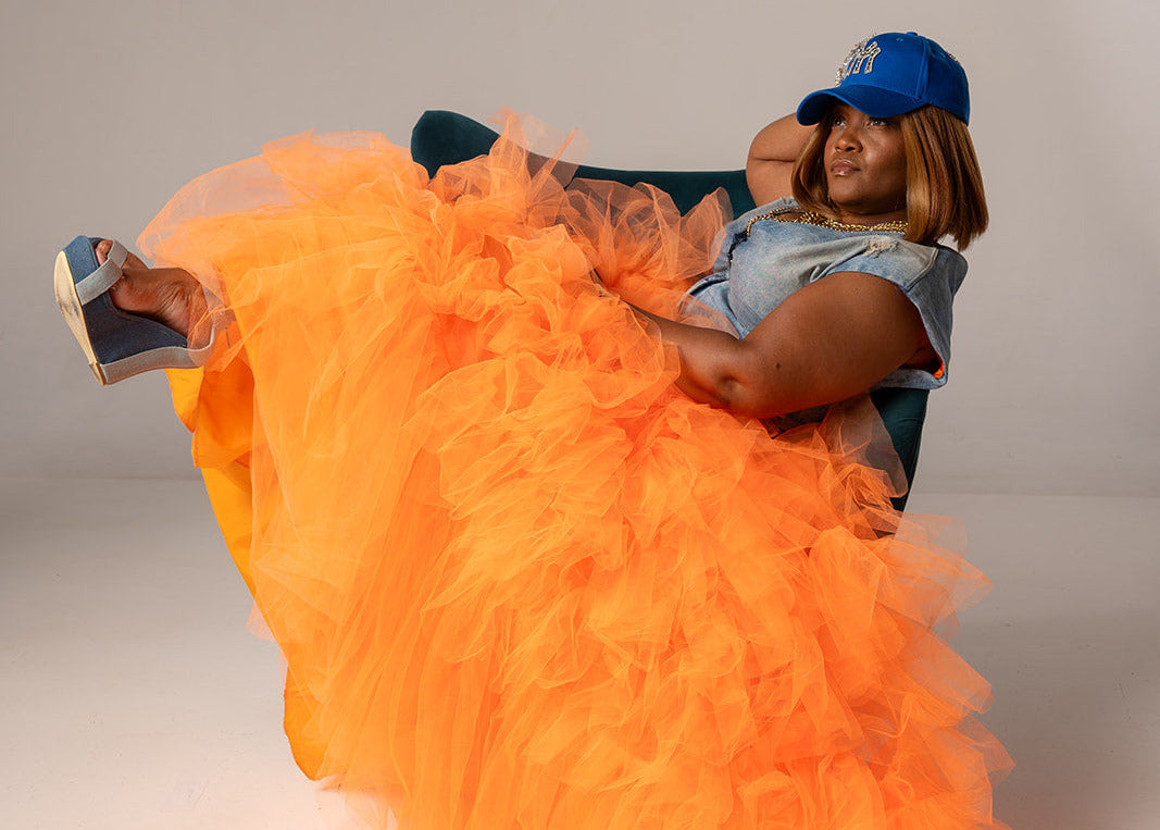 A woman reclining on a dark chair wearing a voluminous bright orange tulle skirt, a light blue denim top, and a blue baseball cap against a plain light gray studio background.